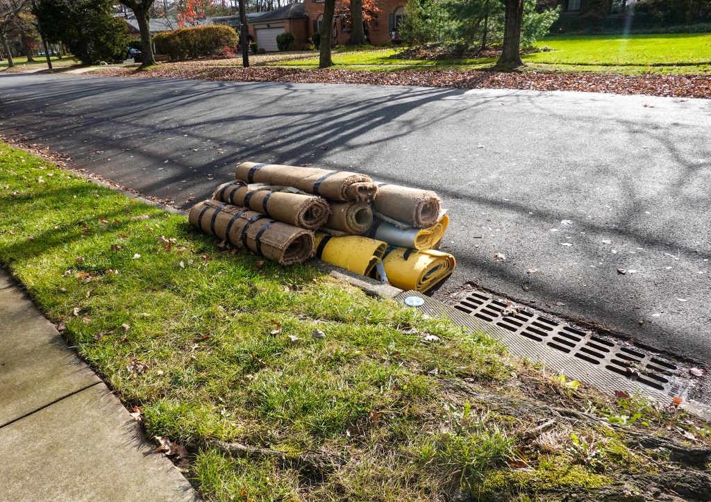 The image shows several rolls of old carpet and flooring materials piled at the curb on a residential street, likely set out for disposal or pickup.RetryClaude can make mistakes. Please double-check responses.