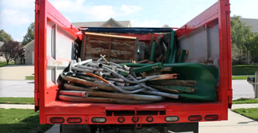 The image shows the back of a red truck or trailer loaded with what appears to be old carpeting, padding, and other demolition or renovation debris in a residential neighborhood.Claude can make mistakes. Please double-check responses.