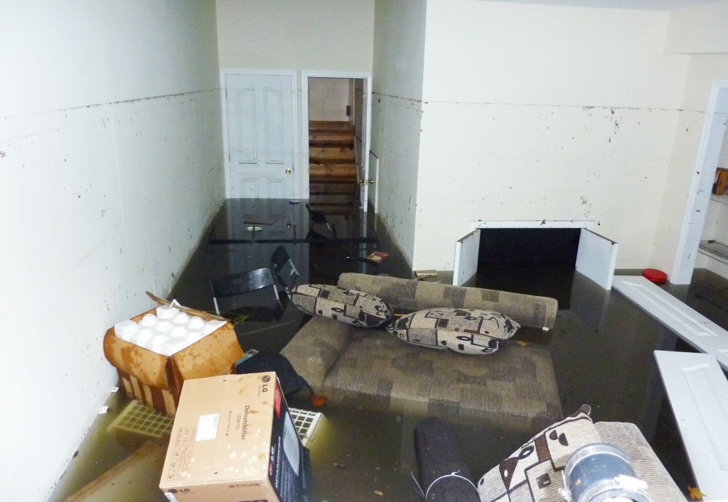 The image shows a flooded basement living room with dark water covering the floor, a soaked couch with pillows, scattered belongings, and water damage visible on the white walls.RetryClaude can make mistakes. Please double-check responses.